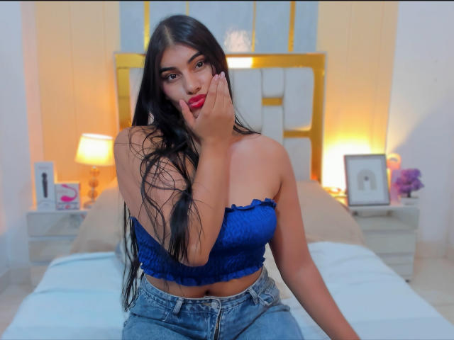 AlaraKey with long black hair, wearing a blue strapless top, blue jeans, and red lipstick, touches her cheek, showing her tits, in a softly lit bedroom.