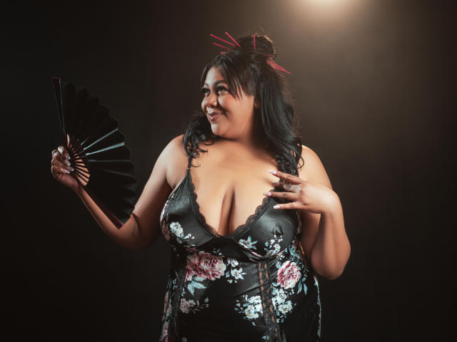 BiancaForest, de talla grande con busto generoso, vestida con un vestido floral negro, sostiene un abanico negro, sonriendo, cabello negro con mechas rojas, fondo oscuro, luz desde arriba..