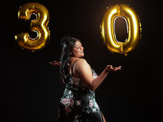 BiancaForest con un trasero grande, vistiendo un vestido floral, cabello negro recogido en un moño, sonriendo, con las manos levantadas, globos dorados con la forma del número "30", fondo oscuro, de pie de lado, luciendo feliz..