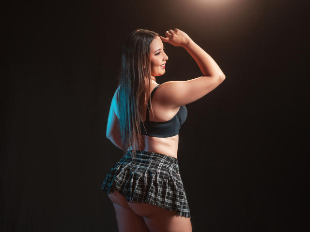 AbellaRiider with long brown hair, black sports bra, and plaid skirt, showing her toned butt, raising her arm, smiling, in a dark studio with blue lighting.