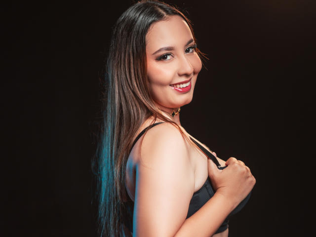AbellaRiider with long brown hair, smiling, wearing a black bra, holding her tits with her right hand, against a dark background. Her red lipstick is noticeable.