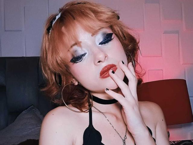 ViolettClay, avec ses cheveux roux, son eye-liner noir, son fard à paupières bleu, faisant la moue avec ses lèvres rouges, un collier ras du cou noir, un soutien-gorge noir et des ongles noirs, touchant ses lèvres avec un doigt..