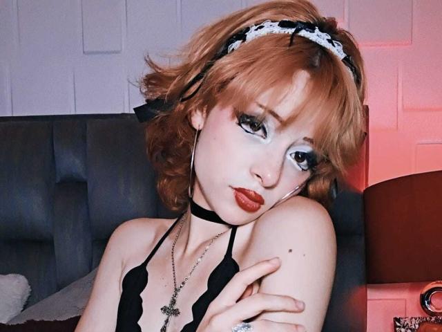 ViolettClay with short red hair, black lace bra, choker, and cross necklace, looking seductively at the camera, arms crossed, wearing white lace headband, red lipstick, and heavy eye makeup.