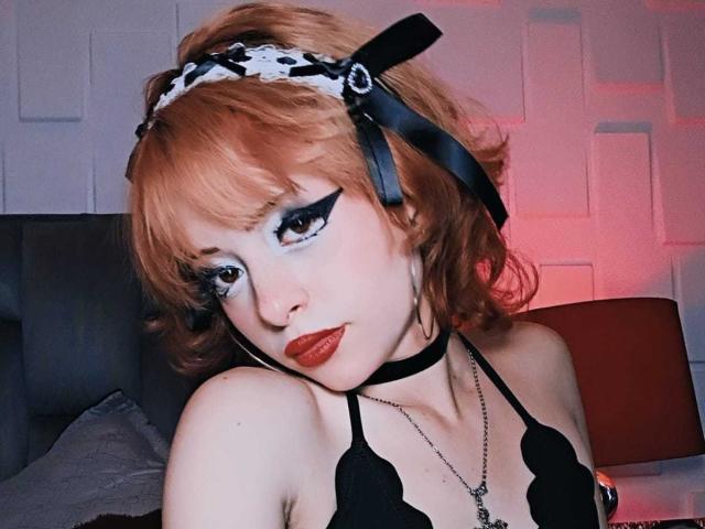 ViolettClay con i capelli arancioni, un reggiseno di pizzo nero, e una fascia per capelli bianca con stampa leopardo, indossa un choker nero, un pesante trucco per gli occhi e rossetto rosso, guardando leggermente verso il basso con un'espressione carina..