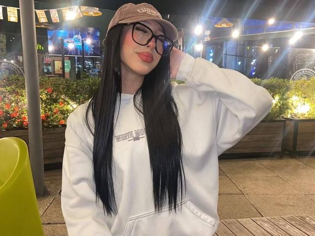 Maddy, con cabello negro largo, gafas y una gorra beige, vestida con una sudadera blanca, sonriendo, con la mano en la gorra, sentada afuera por la noche, una silla verde al fondo..