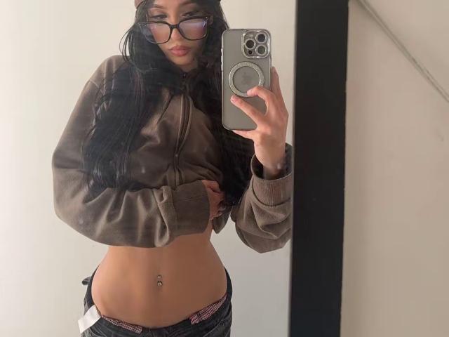 Maddy, portant des lunettes, avec de longs cheveux noirs, une veste marron, laissant apparaître son ventre plat et son piercing au nombril, vêtue d'un jean taille basse, prenant un selfie devant un miroir avec un iPhone..