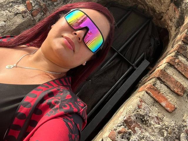 Maya Malik, con i capelli rossi, indossa occhiali da sole riflettenti color arcobaleno, una maglietta nera, una giacca con motivi rossi e neri, una collana d'argento e ha le labbra piene, ed è in piedi vicino a una struttura in mattoni e metallo..