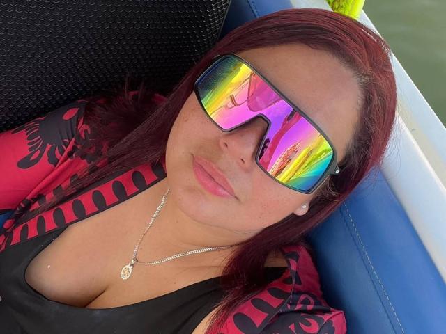 Maya Malik, con i capelli rossi, indossa occhiali da sole riflettenti color arcobaleno, una maglietta nera e una giacca rossa con motivi, mostrando il seno, seduta su una barca blu, con una collana con un ciondolo circolare..