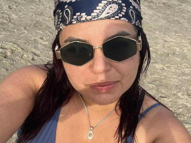 MayaMalik with dark sunglasses, a blue bandana, and a silver necklace, showing her tits in a blue tank top, sitting on a rocky beach.