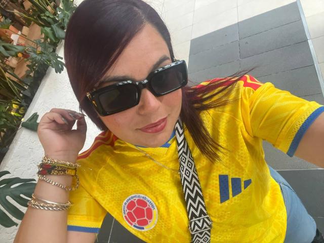 Maya Malik, avec des lunettes de soleil noires, un maillot de foot jaune avec des détails bleus, portant des bracelets, se tenant les cheveux, souriant, montrant son rouge à lèvres rose, et portant une cravate à motifs..