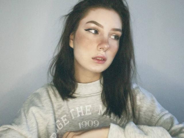 MiraLofi mit dunklen, welligen Haaren, heller Haut und Eyeliner im Winged-Stil, trägt einen grauen "The North"-Pullover, die Arme verschränkt, blickt zur Seite und hat einen leicht schmollenden Gesichtsausdruck..