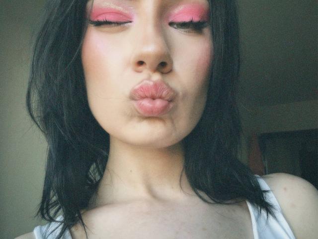 MiraLofi with black hair, pink eyeshadow, and pouty lips, making a kissy face, wearing a white top, in a soft-lit selfie. Her eyelashes are long and curled.