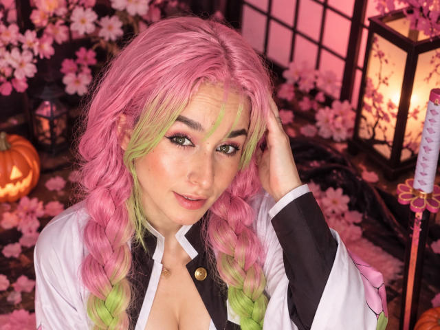 MilaGonz avec des cheveux tressés roses et verts, portant une chemise blanche et noire, une main dans les cheveux, regardant la caméra, avec un eye-liner vert, des fleurs de cerisier roses, une citrouille en arrière-plan..