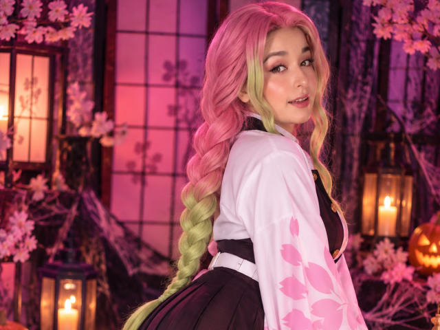 MilaGonz con capelli intrecciati rosa e verdi, che indossa un kimono bianco e nero, sorride alla fotocamera, con luci rosa e viola, lanterne e fiori di ciliegio sullo sfondo..