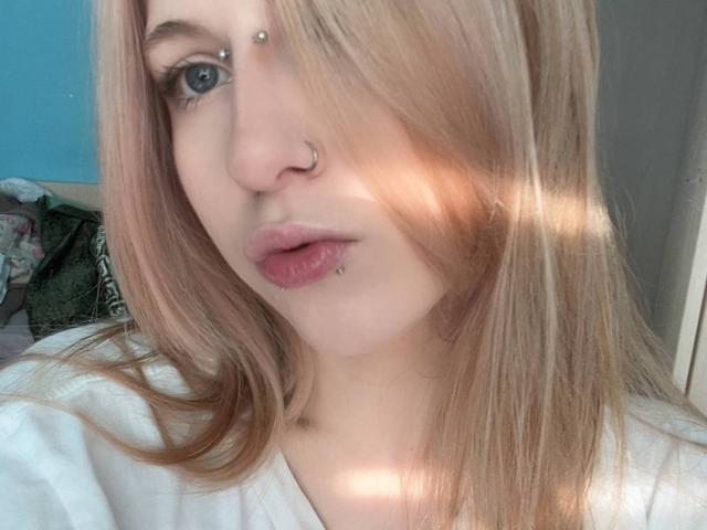 MilaLoner hat blondes Haar mit rosa Schattierungen, blaue Augen, eine Nasenring und eine Lippenpiercing mit kleinen Steinchen. Sie trägt ein weißes Hemd, wobei weiches Sonnenlicht hindurchscheint..
