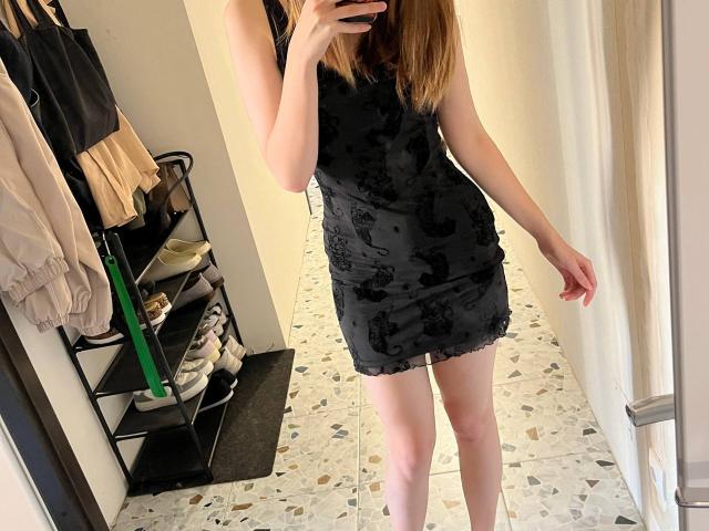 MilaLoner indossa un miniabito nero senza maniche con un motivo in pizzo, è in piedi in un negozio e sta scattando un selfie allo specchio, mostrando i glutei e le gambe, con uno smartphone in mano..