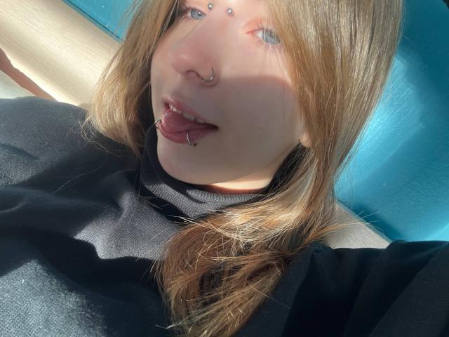 Mila, com cabelo castanho claro, olhos azuis, piercing no nariz, piercing na língua e piercing no lábio, língua de fora, vestindo uma blusa preta, com luz do sol no rosto..