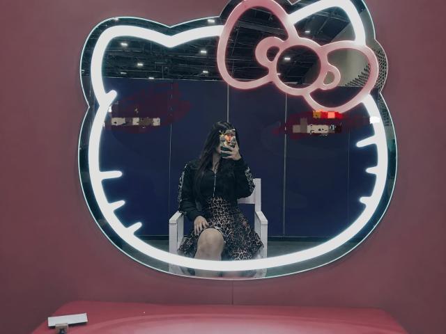MiluSantoro with long black hair, wearing a black top and leopard print dress, sitting in front of a glowing pink Hello Kitty mirror, holding a phone with a cat face case.