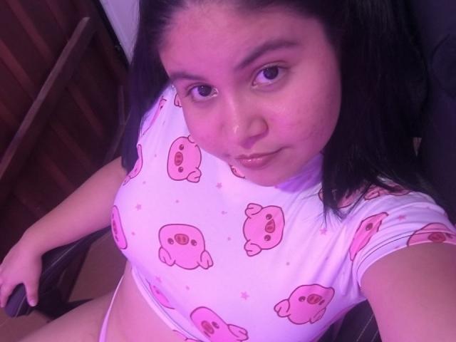 Kappuru with pink bear print crop top, sitting, showing tits, brown eyes, black hair, slight smile, holding remote, purple lighting, cute, chubby, no visible ass or pussy.