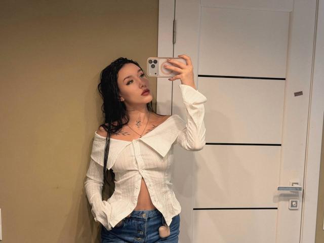 NikkiGoldX with wet black hair, white off-shoulder shirt, and blue jeans, holding a phone, showing a small tattoo on her neck, standing in front of a white door.