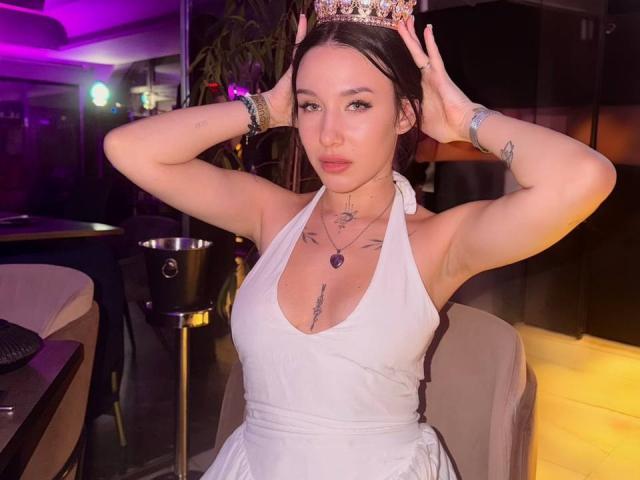 NikkiGoldX with hands on head, wearing a white halter dress, crown, and jewelry, showing small tits and tattoos, in a dimly lit bar with purple and yellow lights.