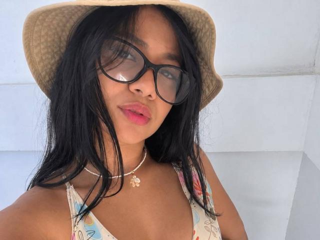 AlanaLuxe has dark, straight hair, black glasses, pink lips, tan skin, wears a beige bucket hat, floral top, and a necklace with a small pendant, showing her tits.