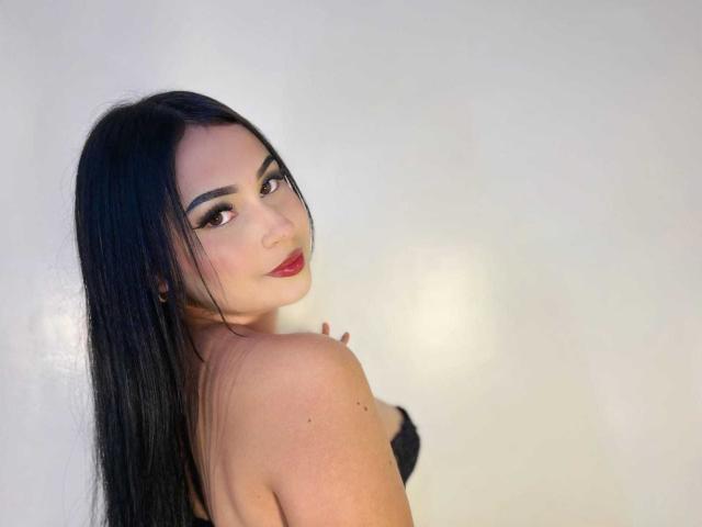 HannahBecker with long black hair, looking over her shoulder, wearing black lingerie, red lipstick, and makeup, showing her bare shoulder and tits. She's smiling slightly.