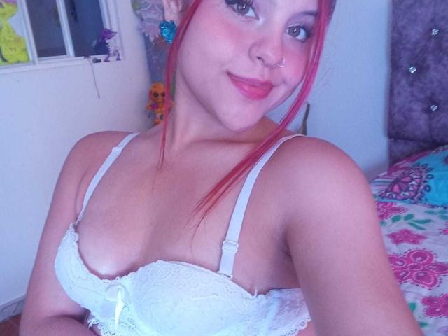 SophieHill with red hair, white lace bra, small tits, cute nose ring, smiling, pink lipstick, blue earrings, bed with colorful patterned blanket, soft light.