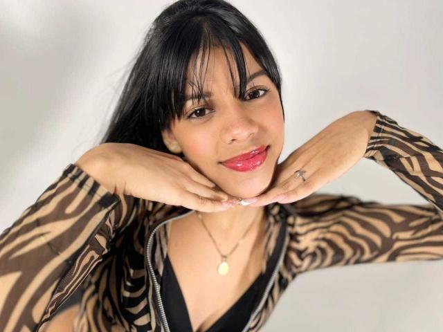 JeanneLacroix with dark hair, brown zebra-pattern top, hands on cheeks, black cleavage, red lipstick, smiling, ring on right hand, necklace with small pendant.