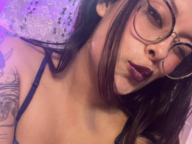 GabyJoy, avec du rouge à lèvres foncé, des lunettes et un piercing au nez, montrant des tatouages sur son bras droit, portant un soutien-gorge noir, et regardant l'appareil photo avec des lèvres pincées..