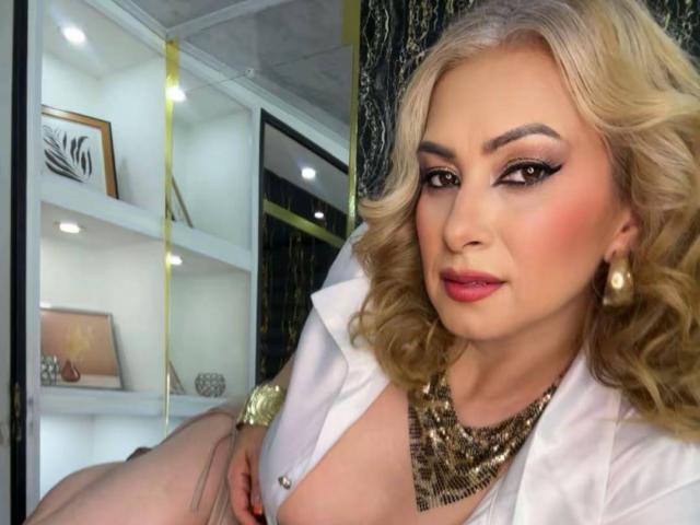 MisseScarleth with blonde curls, red lipstick, and heavy eye makeup, wearing a white shirt and gold necklace, showing her boobs in a mirror selfie.