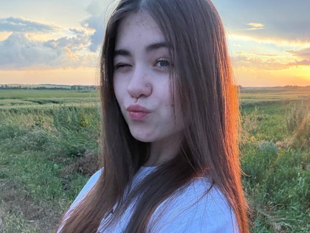 LeliaSixon, con el pelo largo y castaño, guiñando un ojo, frunciendo sus labios rosados, vestida con una camisa blanca, de pie en un campo cubierto de hierba durante el atardecer, con un cielo naranja y azul..