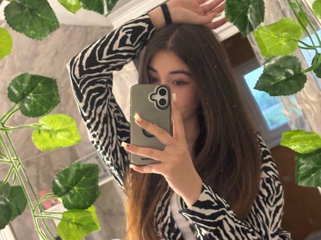 LeliaSixon, avec de longs cheveux bruns, portant un haut à motifs zébra, tenant un téléphone devant des feuilles vertes, prenant un selfie devant un miroir avec un bras levé..