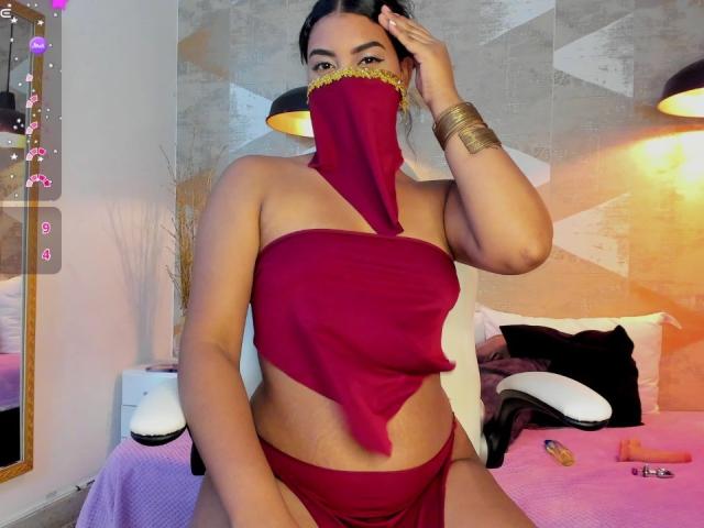 Lalila: Een bruinharige vrouw met een medium bruine huid, donker haar, die een rode maskers draagt, een rood topje zonder bandjes en rode broek, een gouden armband, zittend op een bed met een roze deken, een dildo en een triller..