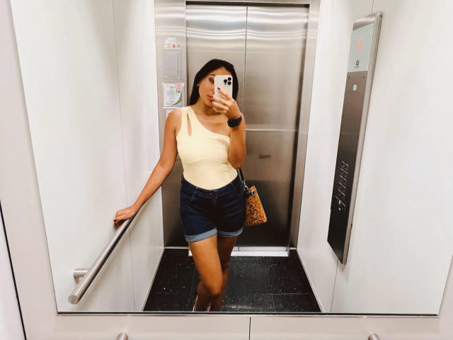 AmeliRoux, with tan skin and long black hair, stands in a elevator, wearing a yellow one-shoulder top, denim shorts, and a leopard print purse, holding a phone.