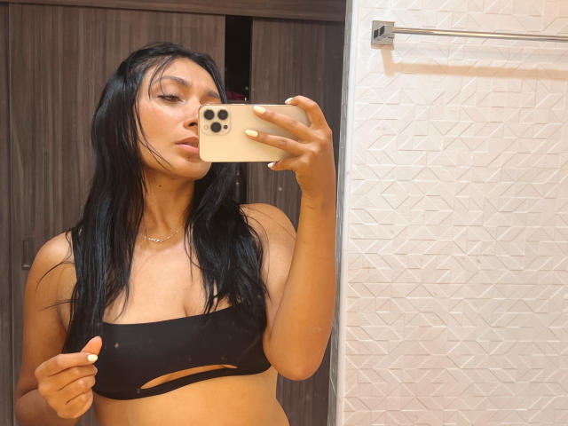 AmeliRoux with long black hair, tan skin, and small tits in a black cut-out top, taking a mirror selfie with a beige phone, in a bathroom.