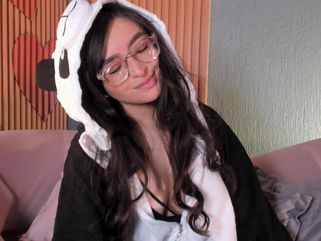 EvaMcAllister, usando óculos, com cabelos castanhos longos e ondulados, vestindo um moletom preto e branco com a estampa de um rosto de animal fofo, com um leve sorriso, mostrando os seios através de uma blusa com decote baixo..