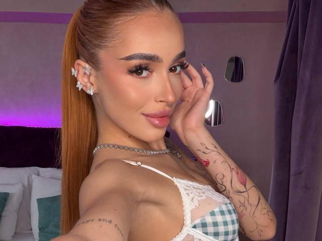MarissaFex, con lunghi capelli castani raccolti in una coda di cavallo, indossa un reggiseno a quadretti con pizzo, orecchini a diamante e una collana choker, mostrando tatuaggi e labbra carnose, mentre si tocca il viso..