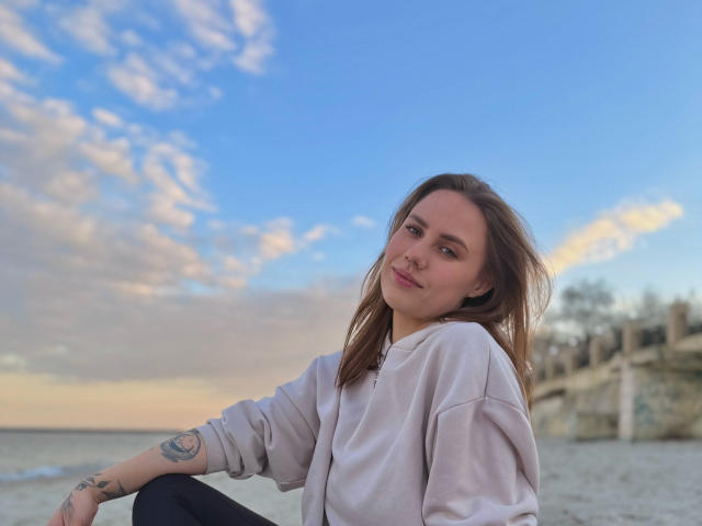 BettyLuv with light brown hair, smiling, wearing a white sweatshirt, sitting on a beach, tattoo on right arm, blue sky with clouds, and gentle ocean in the background.