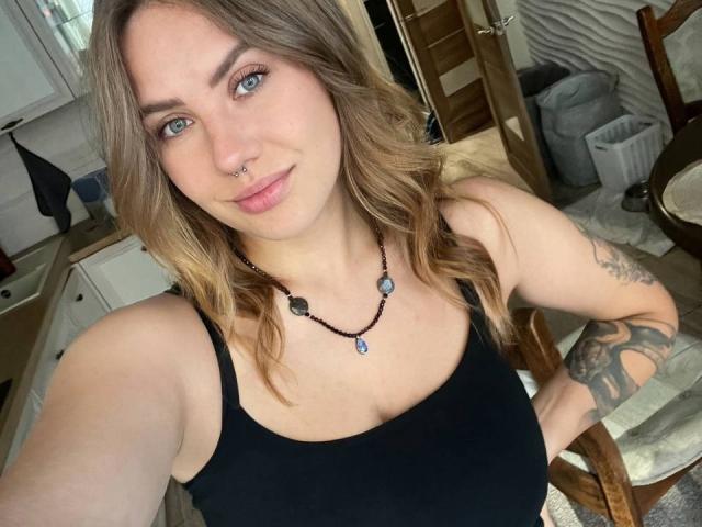 BettyLuv with wavy blonde hair, blue eyes, nose ring, black tank top, necklace with blue gem, tattoo on right arm, smiling, selfie in kitchen. Boobs visible.