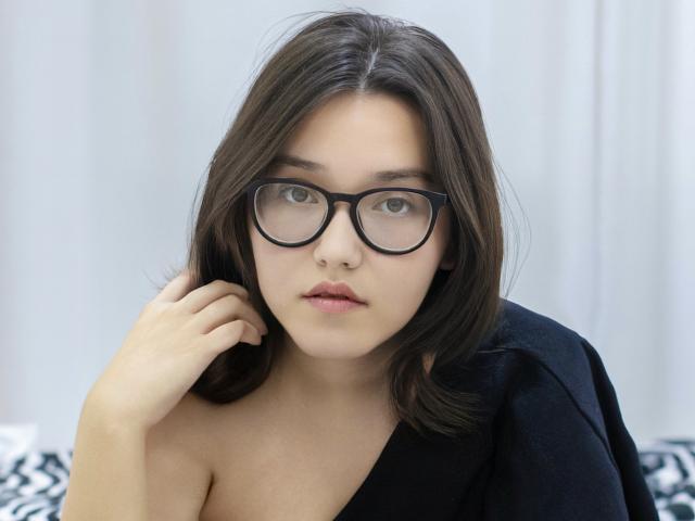 Lily Sinclair, usando óculos, com cabelo castanho de comprimento médio, vestindo uma blusa preta e com uma expressão séria, segurando o cabelo perto do rosto. Seus seios estão parcialmente visíveis. Ela está olhando para a câmera..
