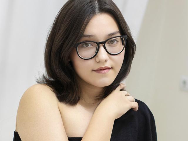 Lily Sinclair, avec ses cheveux bruns moyens, ses lunettes et son haut noir sans bretelles, se tient la joue, a un joli visage et de petites seins, et regarde l'appareil photo avec un léger sourire..
