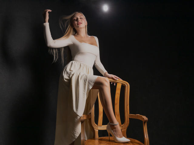Nancy Brooks, com seus longos cabelos loiros, vestido branco e saltos, está em pé com uma perna em cima de uma cadeira de madeira, a mão apoiada na parede, com uma expressão confiante e um leve sorriso..