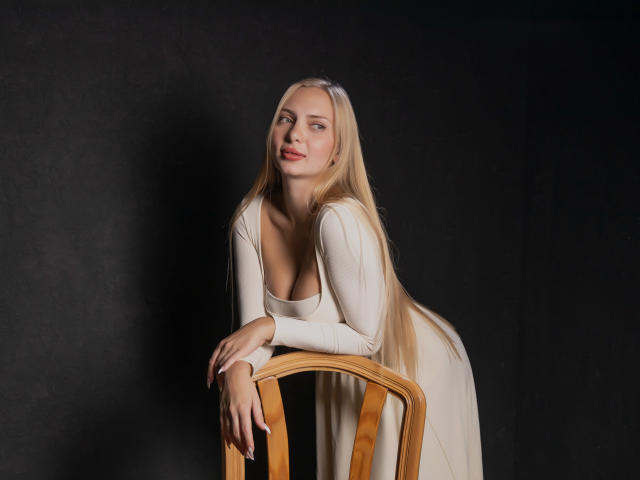 Nancy Brooks, con lunghi capelli biondi, appoggiata a una sedia di legno, indossa un abito bianco, mostrando il seno, su uno sfondo scuro, e guarda leggermente di lato..