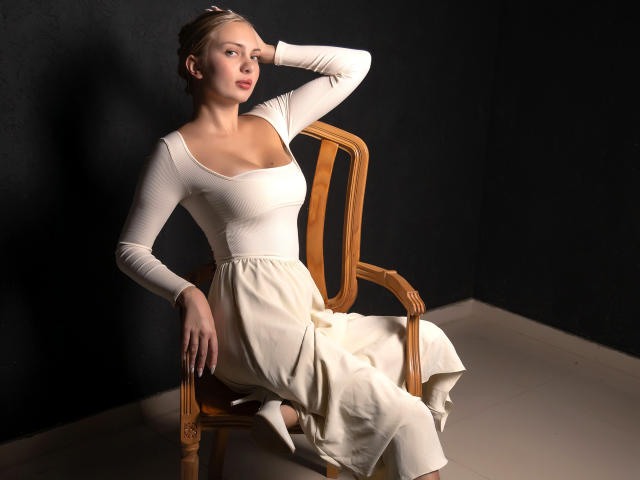 Nancy Brooks, con senos pequeños, vestida con un vestido blanco y una diadema, está sentada en una silla de madera, con una mano en la cabeza, en una habitación oscura, mirando ligeramente hacia un lado..
