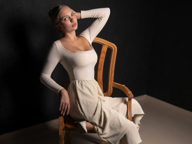 Nancy Brooks, con senos pequeños, vestida con una camiseta blanca ajustada de manga larga y una falda color crema, está sentada en una silla de madera, con la mano detrás de la cabeza, en una iluminación tenue, mirando hacia un lado..