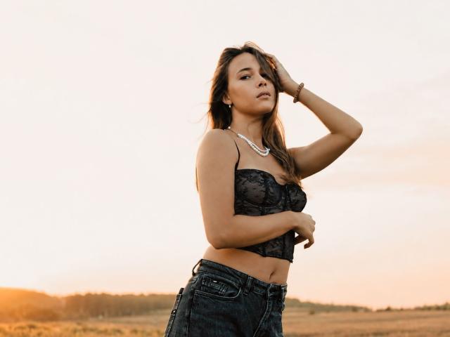 AmmaLoyXX with tanned skin, brown hair, wearing a black lace bralette, pearl necklace, and dark jeans, hand in hair, standing in a field at sunset, looking confident.