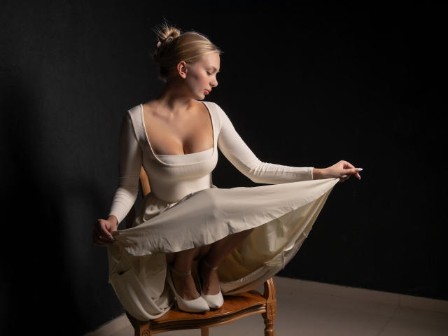 Nancy Brooks mit großen Brüsten in einem weißen Kleid, das sie vor sich hält, sitzend auf einem hölzernen Stuhl, weiße Schuhe, in einem dunklen Raum, blondes Haar zu einem Dutt gebunden..
