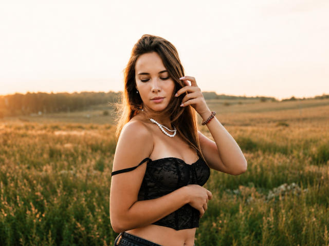 AmmaLoyXX with brown hair, wearing a black lace bra, necklace, and bracelet, eyes closed, touching hair, in a sunlit field, showing her tits, with a calm expression.