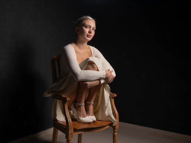 Nancy Brooks, zittend op een houten stoel, heeft kleine borsten, draagt een witte jurk en witte hakken, met haar benen gekruist, in een donkere, minimalistische studio omgeving..